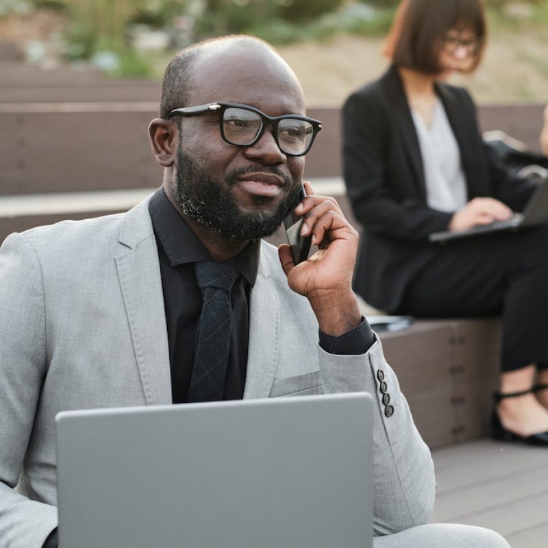 Vertical medium portrait of handsome bearded African American man wearing stylish suit sitting on bench outdoors with laptop talking on phone with colleague